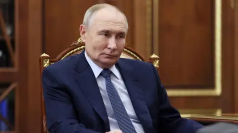 Russian President Vladimir Putin listens during a meeting, wearing blue suit and sat in chair