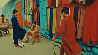 A boy walks down an Afghanistan street rolling a metal wheel - a second boy sits while another stands with his foot on a football