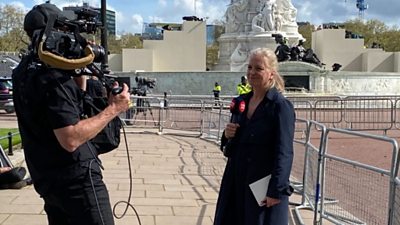 A news reporter on the pavement outside Buckingham Palace, reporting into a camera held by a camera operator.
