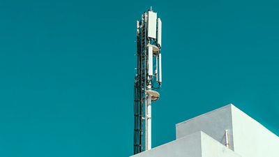 A mobile phone tower above part of a rooftop, set against a clear blue sky