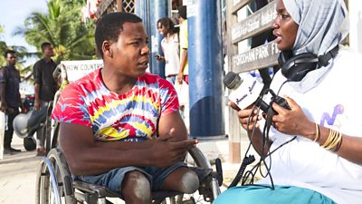 A man, wearing a pink, yellow and white tie dye t-shirt is sitting in a wheelchair, he is being interviewed by a presenter from Niambie radio show. She is wearing a branded t-shirt and headscarf, holding a Niambie microphone