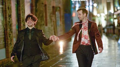Two men jovially walk down a street at night holding hands