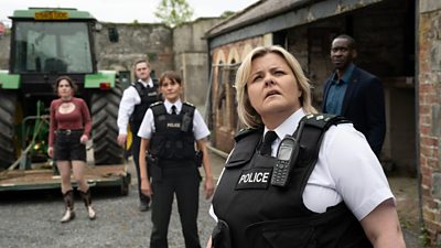 Three uniformed police officers and a man in a suit stand in a yard examining something that is out of view