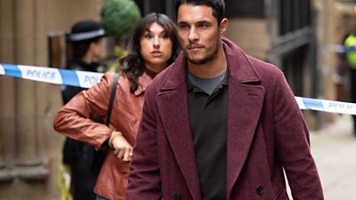 A man walks purposefully in a red coat. A woman follows behind him. There is a police cordon behind her.					