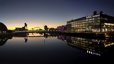 The ѿý Scotland building at Pacific Quay, Glasgow, at dusk. The building and a nearby bridge are reflected in the water that surrounds the building.	