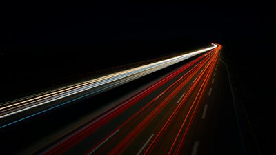 An abstract image of some lights (presumably from a car or traffic) blurred across the frame as they moved too fast for the shutter, creating multiple thin and brightly lit red and white lines on a very dark black background.