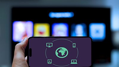A mobile phone being held by a hand in the foreground, showing a graphic of earth, with icons of devices playing iPlayer on them.  Blurred in the background is a large TV screen showing something indistinct.