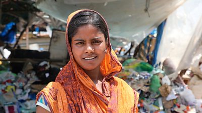 a young woman in india faces the camera - behind her is a large pile of recycling