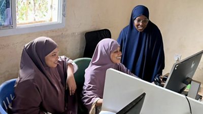 Three Somali journalists are working around a computer, one wears a blue headscarf and glasses, the other two are wearing different shades of purple.