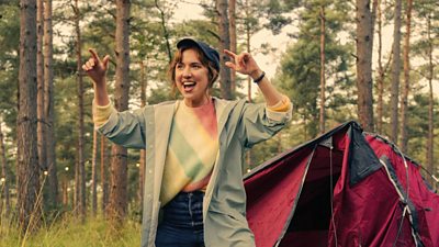 Nikki (Esther Smith) poses with her hands in the air pointing in front of a half assembled tent in a forest. Nikki wears jeans, a multicoloured jumper, boots and a grey coat.