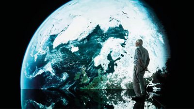 David Attenborough stands on a black reflective floor looking at a screen with a giant projection of planet earth.