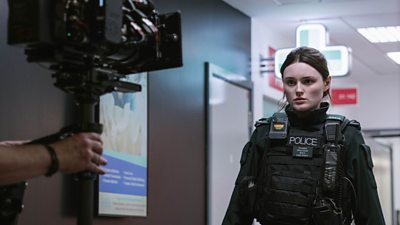 Annie (Katherine Devlin) stands in front of a camera. She is dressed in police uniform. She has brown hair tied up and a serious expression.