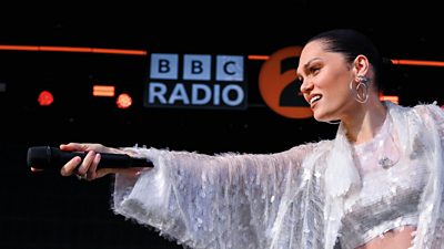 Jessie J stands on stage in front of BBC Radio 2 branding. She is wearing a matching white sparkly outfit and holds the microphone to the audience, encouraging them to sing.