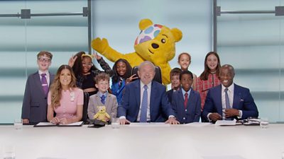 Lord Alan Sugar sits in The Apprentice boardroom smiling, surrounded by child contestants of Mini Apprentice, all wearing suits. Pudsey stands behind the lineup waving.