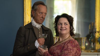 Mr. and Mrs. Bennet (Richard E. Grant and Ruth Jones) stand in large, open room. Both look to the right, Mr Bennet with a scowl and Mrs Bennet with a smile. Both hold wine glasses. Mr Bennet wears small wired spectacles and period dress, and Mrs Bennet wears a period pink floral dress and a gold necklace.