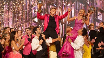 Winners Witney Carson and Robert Irwin are lifted onto cast members shoulders as they lift the glitterball thropy. They are on a stage in front of dancing with the stars branding and Witney wears a pink sparkly dress and Robert wears a pink jacket and black trousers and shirt.