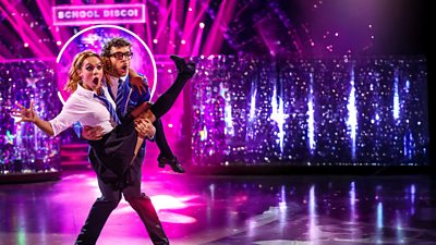 George Clarke and Alexis Warr dance the Charleston. Both wear matching school uniform and George lifts Alexis into the air. Both have shocked expressions.