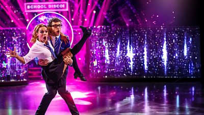 George Clarke and Alexis Warr dance the Charleston. Both wear matching school uniform and George lifts Alexis into the air. Both have shocked expressions.