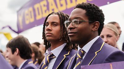Toby (Sekou Diaby) and Omar (Myles Kamwendo) stand sideways in purple school uniform in front of a school banner. Toby has round glasses on.