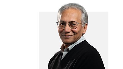 BBC Chairman Samir Shah smiling side on in a black cardigan, striped shirt and black rimmed round glasses.