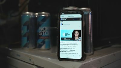 A mobile phone on a table beside two drink tins - screen shows a woman and the Suspilne logo