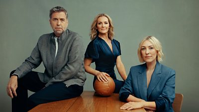 A portrait picture of the three presenters sat on or at a wooden table. Gabby has her hand on an old fashioned leather football. They are all posing for the camera against a grey background.