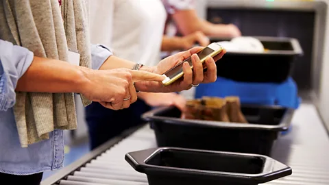 A traveller holds a smartphone while placing belongings into a tray at an airport security checkpoint (Credit: Getty Images)