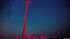 A wind turbine lit up bright red is seen in contrast to a starry sky with the sweeping trail of a meteor in view