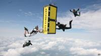 A yellow box falls through the clouds surrounded by skydivers in black costume.