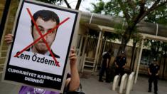 woman holding sign showing Cem Ozdemir's face with a big red X through it, security guards in the background