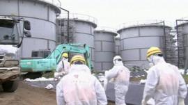 Workers at the plant wearing radiological protective suits, hard hats and masks walking towards large containers