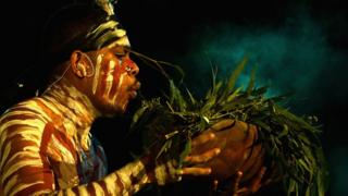 A dancer in Aboriginal dress blows smoke from a bowl filled with leaves.