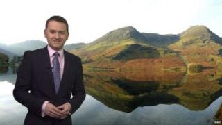 BBC Weather presenter Matt Taylor stands in front of a photo of hills on a cloudy day