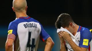 A USA footballer holds his jersey to his face while his teammate looks on.