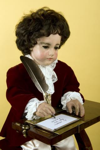 The figures are still working. The boy dips the quill in an inkwell and shakes off the extra ink before writing on the paper.