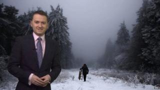 BBC Weather presenter Matt Taylor stands in front of a photo of snow covered trees on a cloudy day