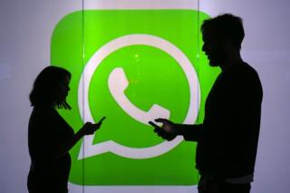 Two people hold their  cell before a poster with the logo WhatsApp. 