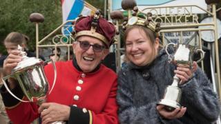 A Chelsea Pensioner and a woman, both wearing crowns decorated with conkers, hold their trophies aloft sitting in chairs that say 'King Conker' and 'Queen Conker'.