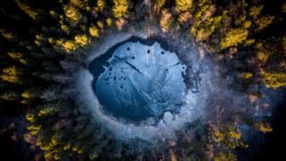 Image shows an aerial shot of a frozen lake surrounded by fir trees