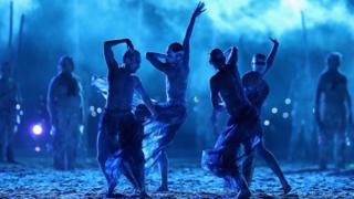 A tribe performs a traditional dance against bright blue light.