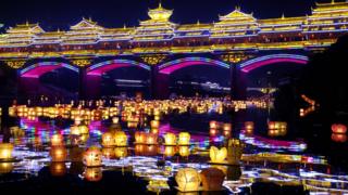 A night scene showing a river covered with hundreds of lanterns, floating under a brightly lit bridge.