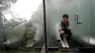 Hombre sentado en un inodoro detrás de una puerta de vidrio ligeramente esmerilado.