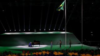The Brazilian flag is raised as musician Carlos Martin, whose right hand is severely disabled, plays the national anthem on the piano.