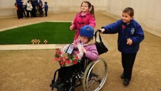 Two children push a third child in wheelchair, carrying a wreath