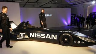 Nissan's new Deltawing race car is seen after it was unveiled in London. The black, sleek sports car has a very long fuselage. The car is unveiled at a press event and people can be seen celebrating, shrouded in purple light.