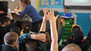 children raising hands in classroom while teacher is turned away