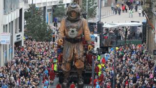 A huge marionette sea diver character walks through the street, surrounded by a crowd of people.