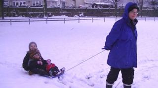 Two girls being pulled along on a sledge in the snow by their mother.
