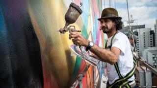 An artist close to the side of the building sprays the wall with an airbrush.