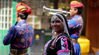 An Indian brass band play in the street, they are wearing brightly coloured clothes and a woman in smiling at the camera